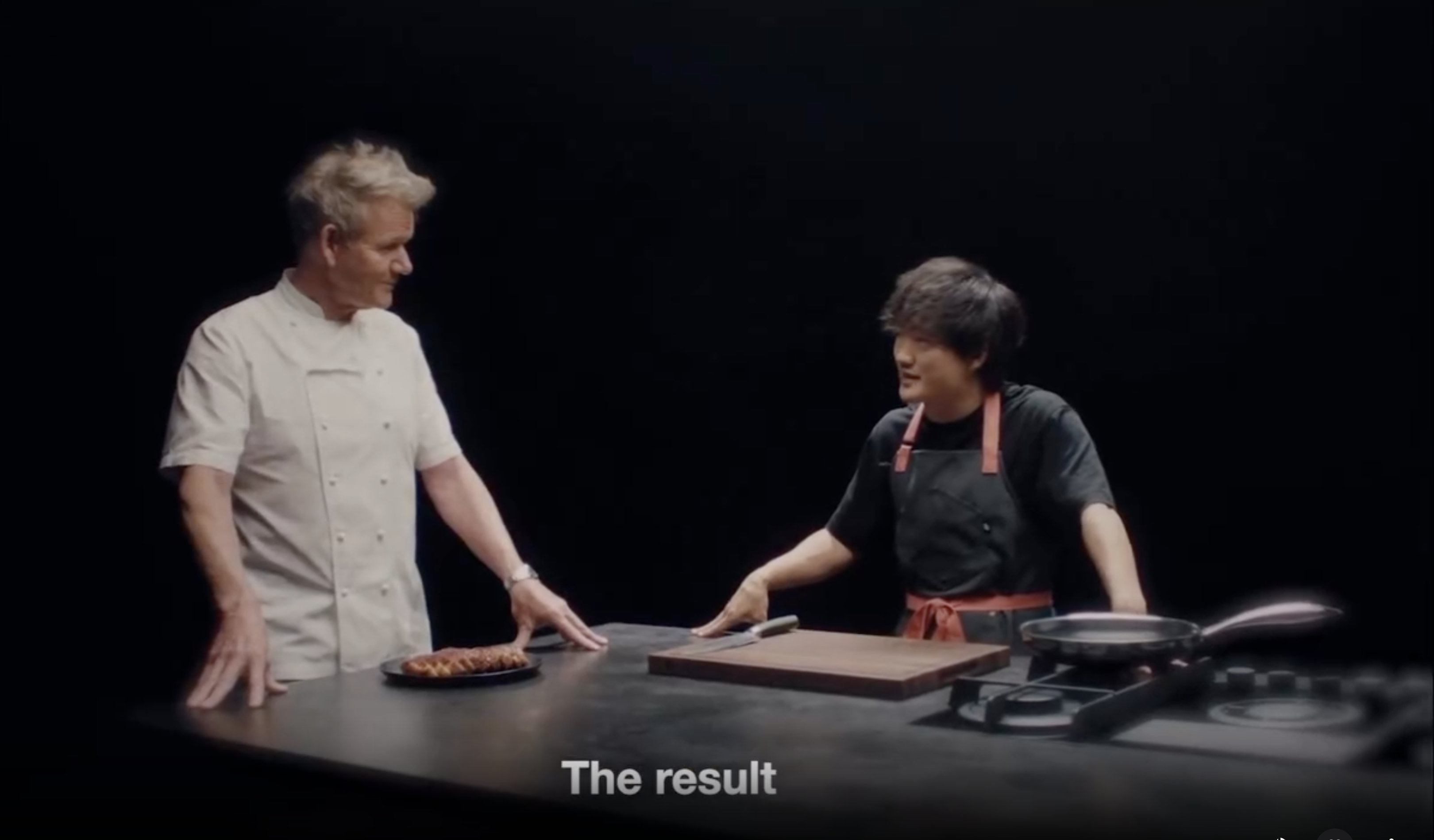 A chef in a white chef's jacket talking with a cook in a black apron across a dark kitchen counter, with the caption The result.