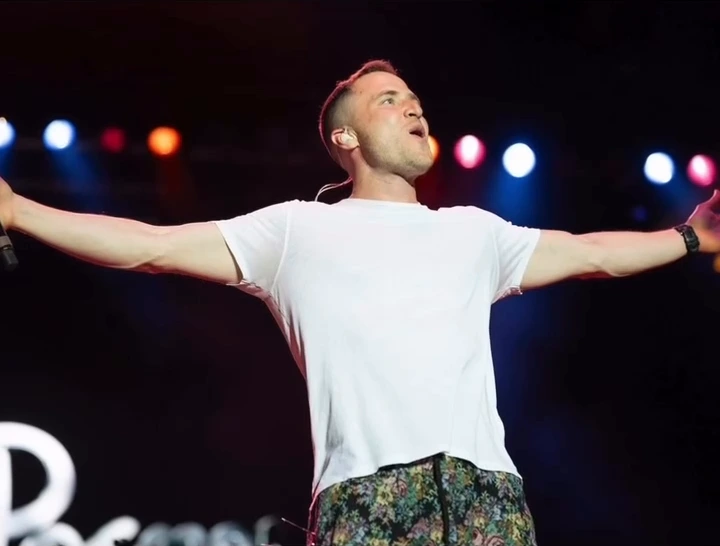 Mike Posner on stage in a white T-shirt with his arms spread wide under colored stage lights.
